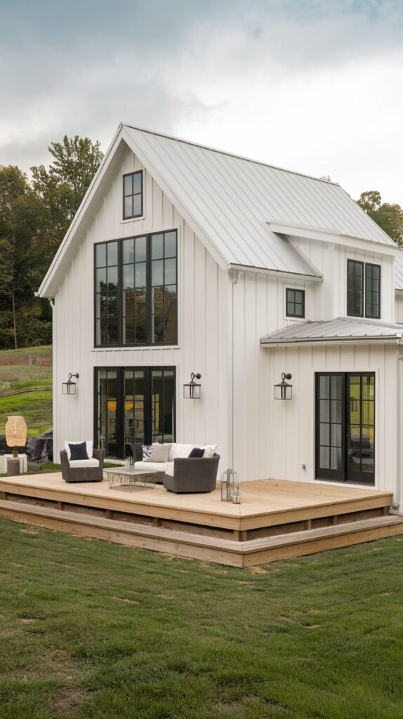 White board-and-batten farmhouse with a light metal roof. The rear facade features a large, low-profile wooden deck accessed by a two-story window wall and a separate set of French doors.