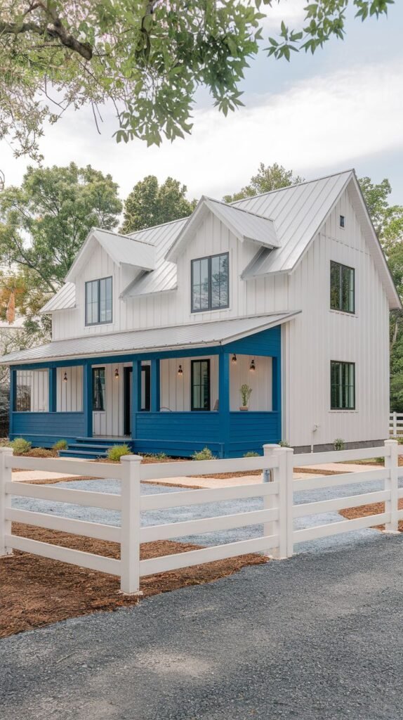Two-story white farmhouse with twin dormers, a light metal roof, and a contrasting bold blue skirting/base wrapping the front porch. The home is framed by a white rail fence and a gravel driveway.