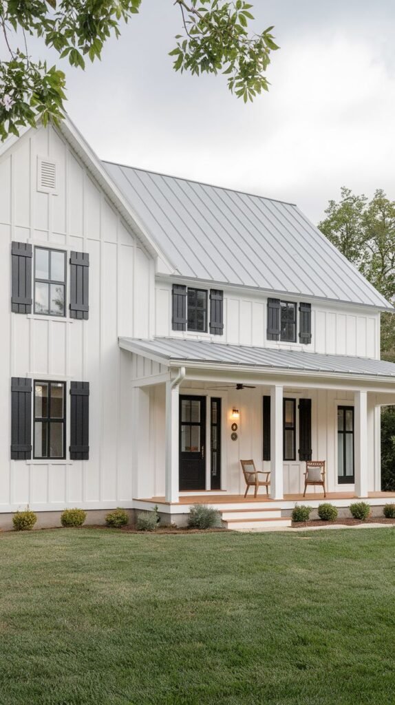 Traditional two-story white farmhouse with a light metal roof and black board-and-batten shutters flanking the windows on both floors. A wide, covered porch spans the front, overlooking a lush green lawn.