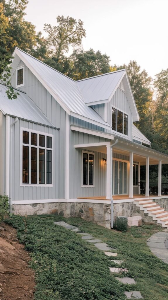 Light blue-green vertical-sided farmhouse with white trim and a grey metal roof. The lower portion rests on a rough-hewn stone foundation. A winding path of stone stepping blocks ascends the grassy slope towards the porch.