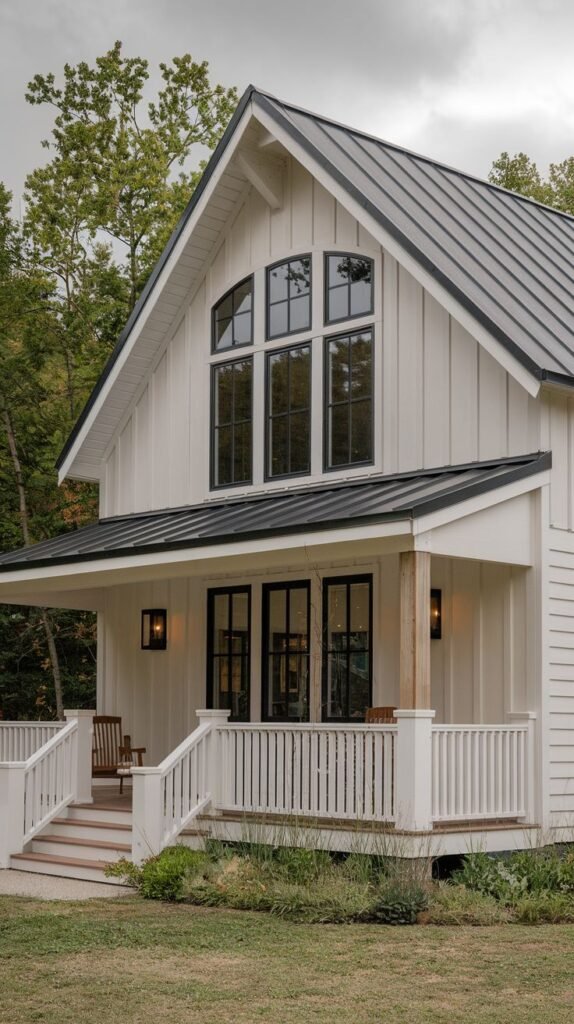 White board-and-batten farmhouse with a dark roof. The gable end features a two-story window configuration topped by a gentle arch. The front porch features traditional white railing and exposed wooden support posts.