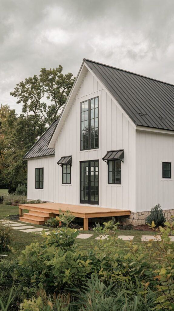 White vertical-sided farmhouse with a steep black metal roof. The central entry consists of tall black-framed glass doors and windows, flanked by smaller windows topped with small black metal awnings.