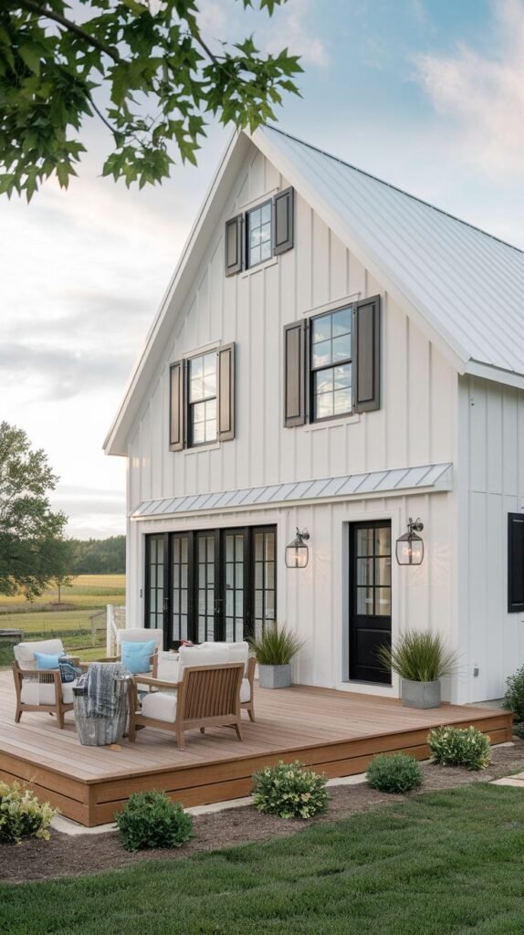 White vertical-sided farmhouse with a gabled roofline, featuring dark traditional shutters flanking windows on the upper level, and a low, expansive wooden deck furnished for outdoor lounging.
