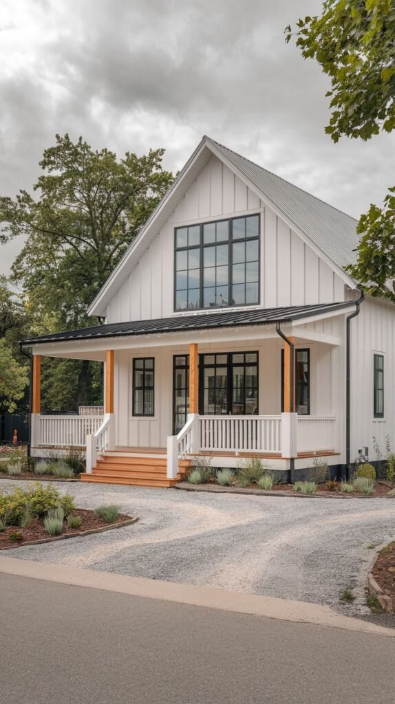 White two-story farmhouse with a central gable and a covered porch. The porch is supported by natural wood posts and framed by white balustrade railing, with warm wooden steps leading down to a gravel driveway.