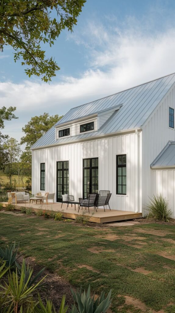 Simple, long white vertical-sided farmhouse with a light silver metal roof and a small shed dormer. A large, uncovered wooden deck extends from the side, accessed by multiple black-framed glass doors and windows.