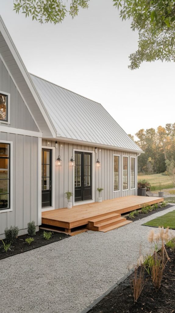 Single-story structure with soft grey vertical siding and a silver metal roof. A long, low natural wood deck provides access through black-framed French doors, approached by a wide gravel path.