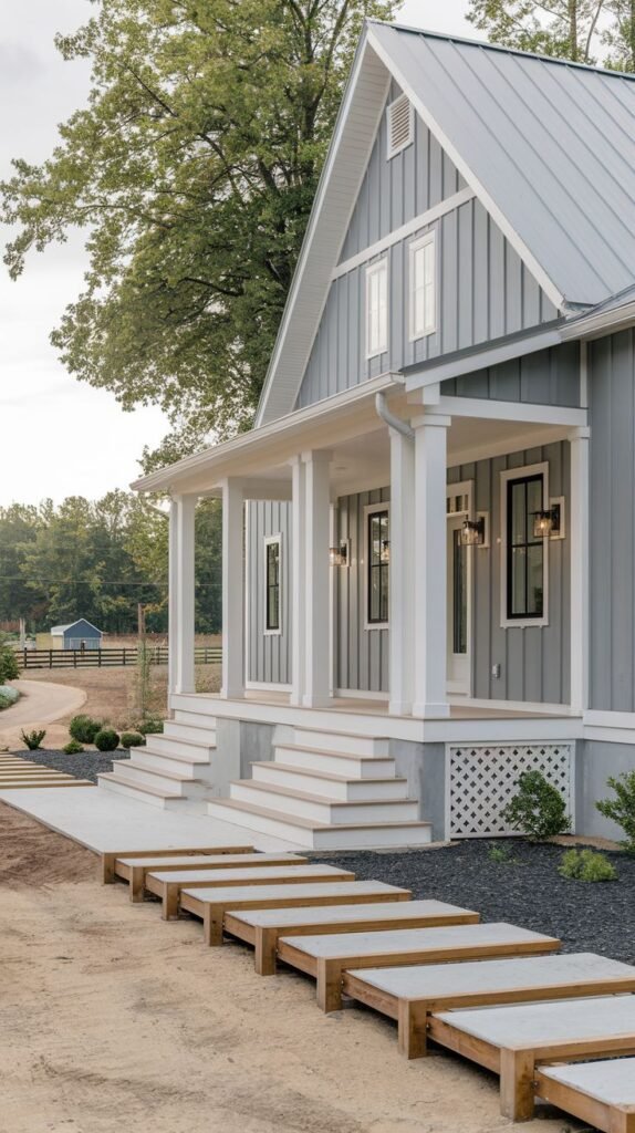 Blue-grey vertical-sided farmhouse with a front porch supported by large white columns. The entryway features contemporary steps composed of large, horizontal wooden planks interspersed with dark gravel.