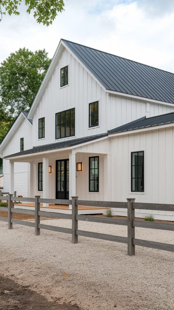 Two-story white farmhouse featuring a dark metal roof and a full-length covered porch. The property entrance is marked by a weathered, grey wooden split-rail fence separating a gravel drive from the house.