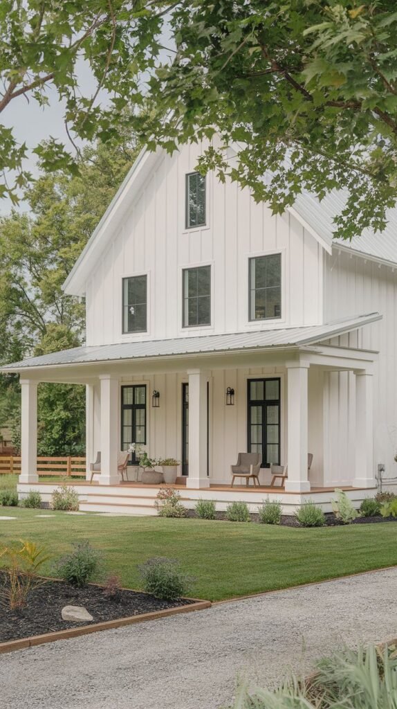 Symmetrical two-story white farmhouse with a light grey metal roof covering a deep front porch. The porch features white columns, wood decking, and two simple chairs, overlooking a manicured lawn and gravel drive.