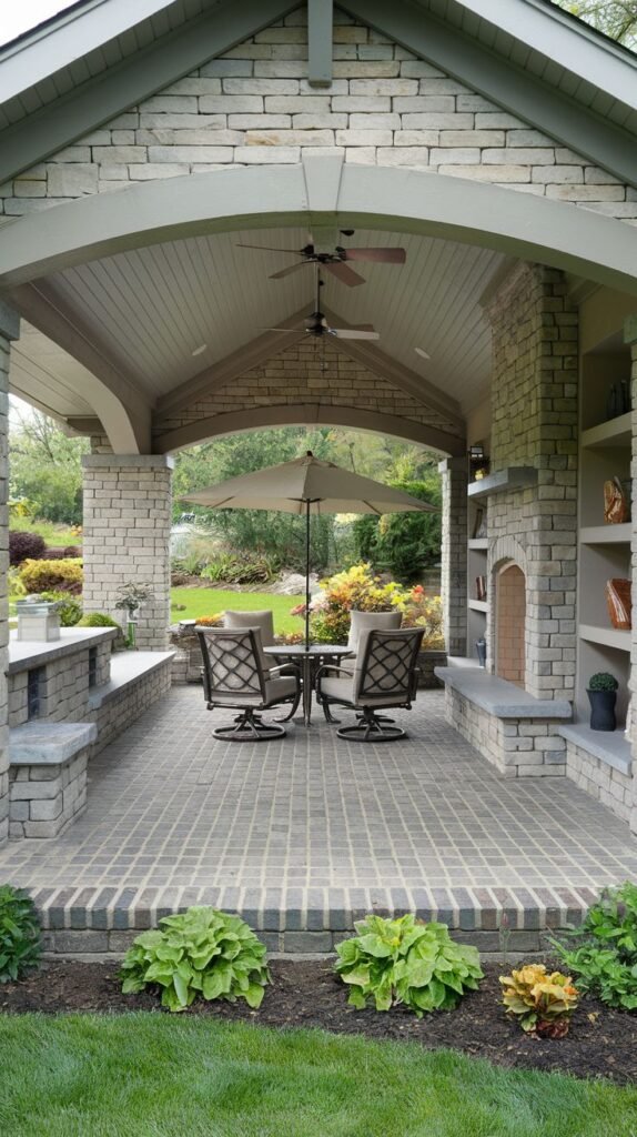 A covered patio with a vaulted ceiling structure. The walls are built of light stacked stone, and the floor uses a dense pattern of dark and light brick pavers. The fireplace is flanked by integrated shelving/niches.