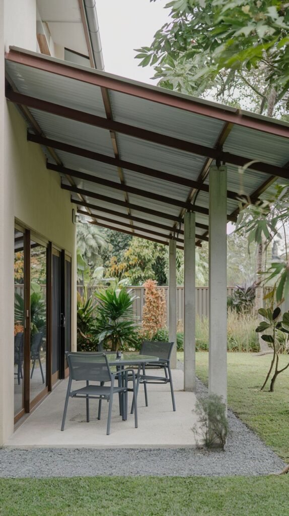 Minimalist patio cover featuring a sloping roof of corrugated metal sheeting supported by widely spaced, rounded concrete columns, shading a small concrete dining area.