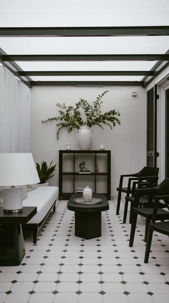 A minimalist black and white patio with patterned geometric floor tiles, black open shelving, and black and white seating under a metal-framed translucent roof.
