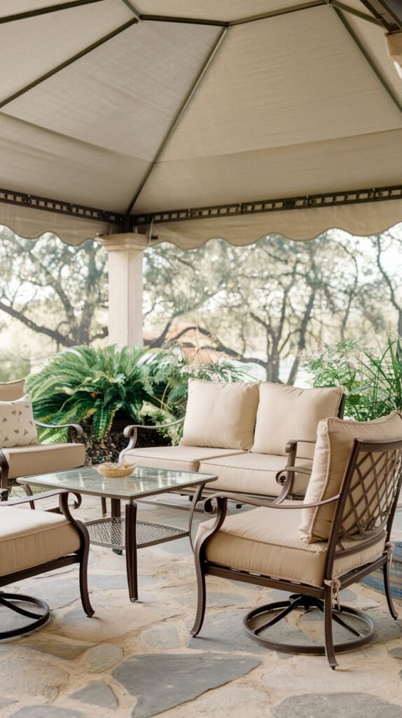 Covered patio featuring brown metal framed outdoor sofa and chairs with beige cushions, clustered around a glass-topped coffee table, set on flagstone flooring under a beige fabric canopy.