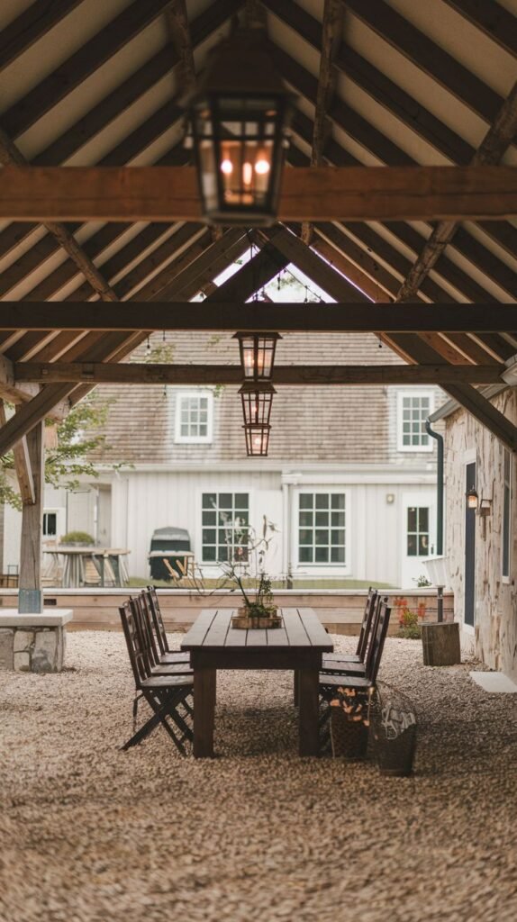 Rustic outdoor dining area beneath heavy timber beams, featuring three traditional dark lantern pendant lights suspended above a wooden table on a gravel floor.