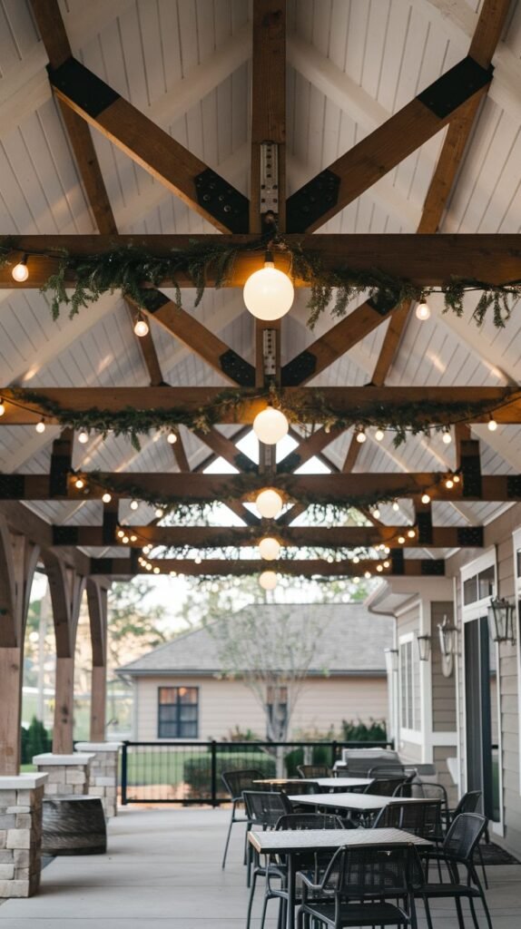 Exposed wooden truss ceiling of a covered patio decorated with string lights (including large globe bulbs) intermingled with festive green garland above multiple outdoor tables.