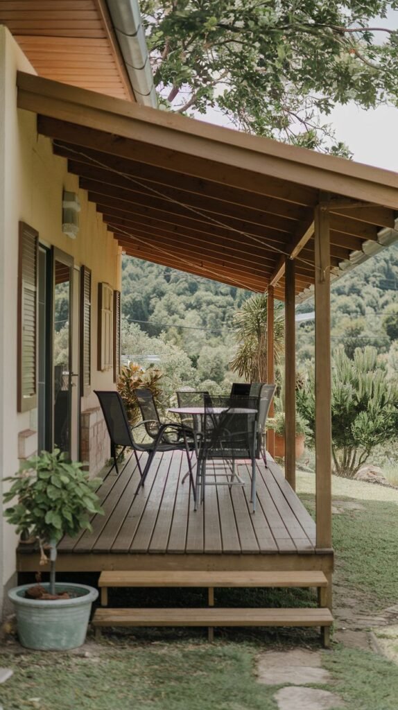 A long, elevated wooden deck attached to a light stucco house, covered by a sloped, dark wood roof structure, featuring a black metal dining set overlooking a lush, wooded valley.