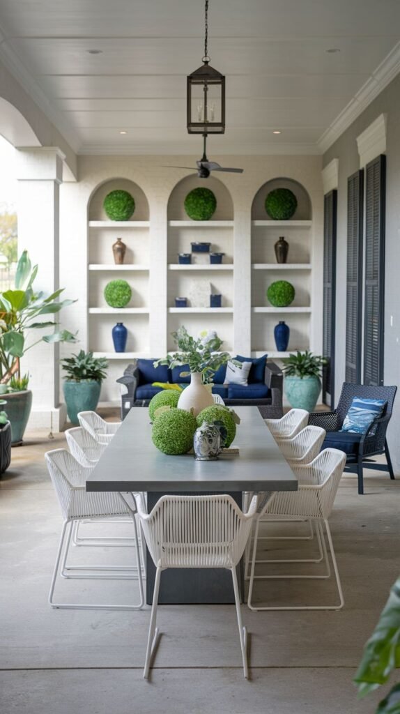 Formal outdoor dining space with a dark gray table and white chairs, backed by built-in white shelving arches displaying green topiary balls and blue vases.