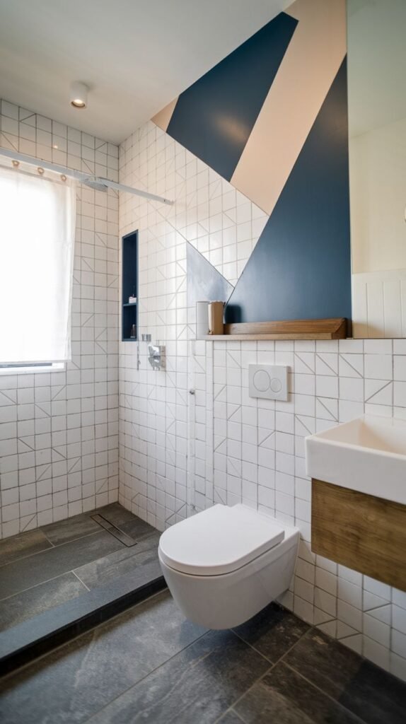 Modern bathroom featuring walls tiled in white geometric grid tiles, accented by a large, angular geometric mural painted in dark blue and pale pink/white, with a floating toilet and dark slate floor.