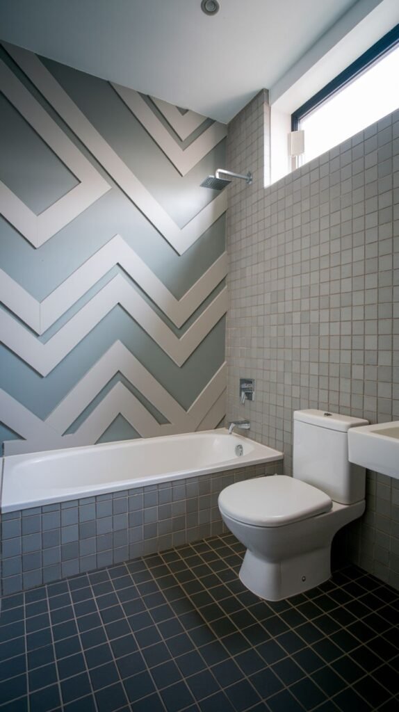 Modern bathroom featuring a layered grey and white dimensional chevron accent panel above a built-in white bathtub, bordered by light grey square tiles on the walls and dark grey square floor tiles.
