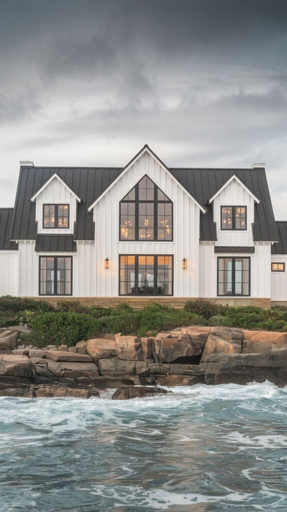 High-contrast black and white farmhouse resting on a rugged, rocky coastal bluff, with ocean waves crashing in the foreground. Features a black metal roof, white board-and-batten siding, and twin dormers flanking a central glazed gable.