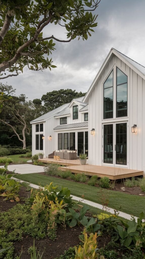 Rear view of a long, white farmhouse structure composed of multiple gables and dormers. A low wooden deck with outdoor furniture sits central to the facade, bordered by a concrete path and garden beds.