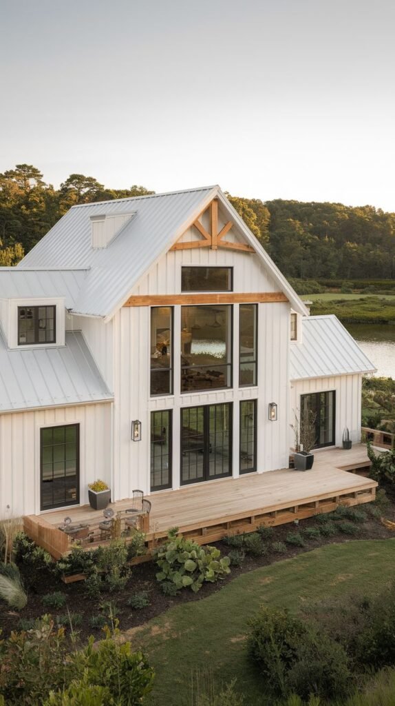 Elevated aerial view of a white waterfront farmhouse with a light metal roof and a prominent exposed natural wood truss detail in the gable. A large light-colored wooden deck extends from the house toward the water.