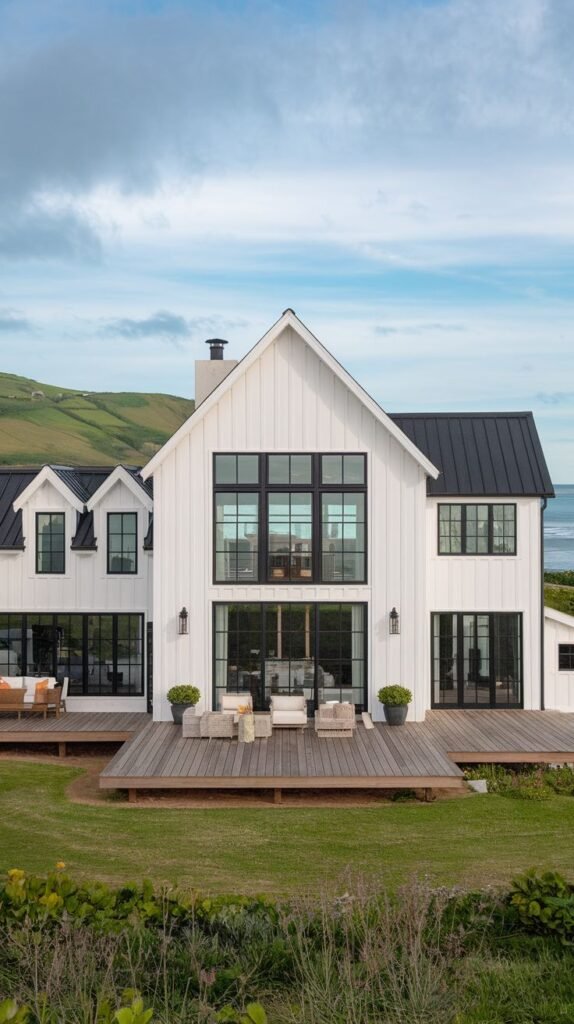 White farmhouse with a black metal roof overlooking coastal rolling hills. The rear facade features a central, tall gable with multi-pane black windows and doors opening onto a wide, low dark wood deck.