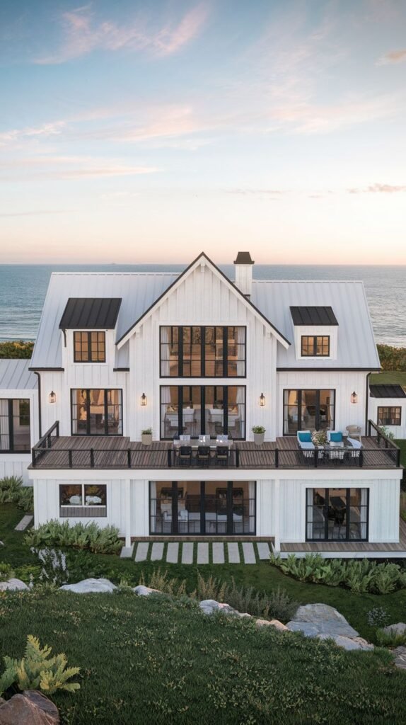 Multi-level white coastal home overlooking the ocean. The structure features black-trimmed dormers and a light metal roof, with an upper dark wood deck and a lower level accessible by glass doors and paver steps.
