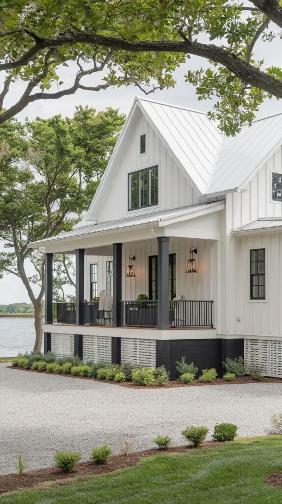 White farmhouse on the water's edge with a white metal roof. A covered porch features bold black columns and railings, contrasting with white lattice skirting below. The approach is a light gravel driveway.