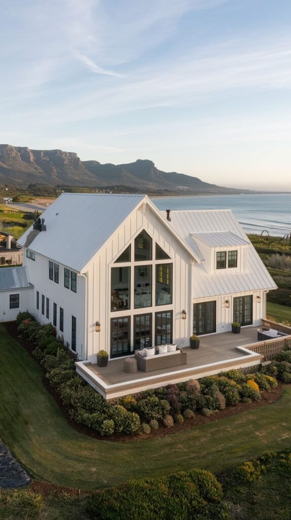 Aerial view of a stunning white coastal farmhouse with a white metal roof, featuring a two-story glass gable facade opening onto a large wooden deck. The house is situated near a beach with mountains in the background.