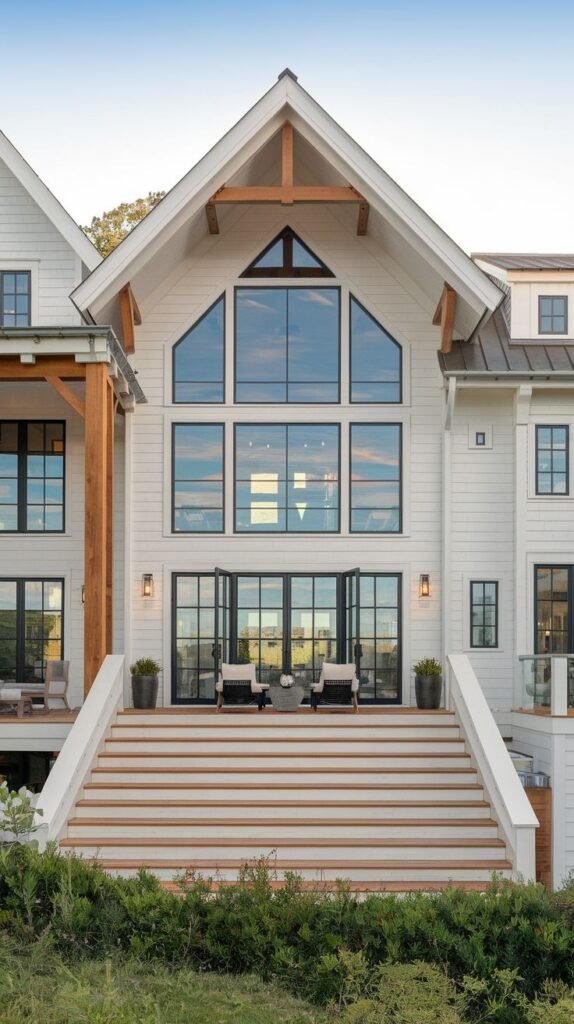 White farmhouse rear view featuring a broad, tiered staircase with white sides and wood treads leading to the main level deck. The central gable displays a large glass wall and an exposed wooden cross-brace detail.