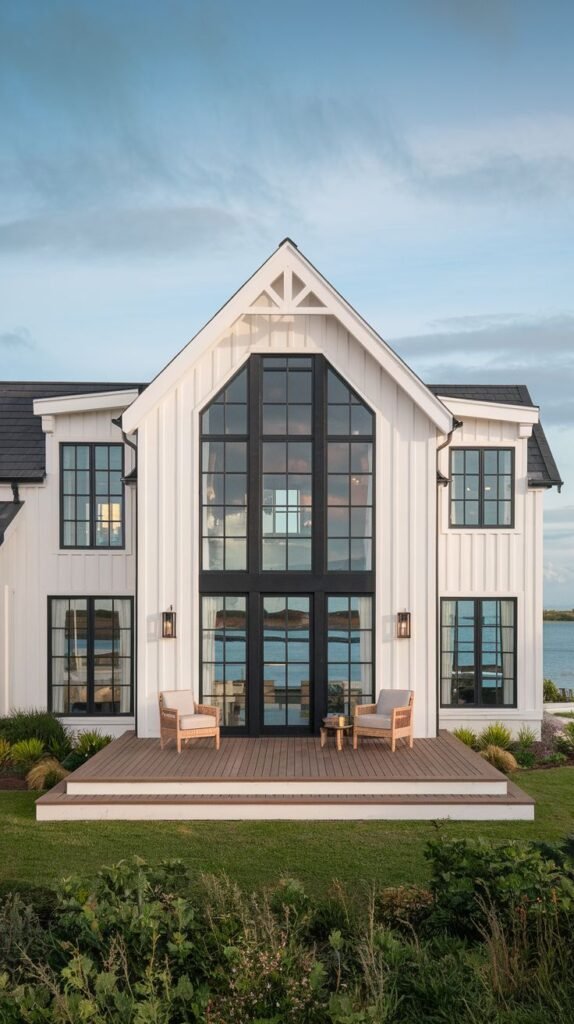 White farmhouse with a high central gable and dark roof, featuring a large black-framed window wall overlooking water in the distance. A small raised deck area sits in front with outdoor chairs.