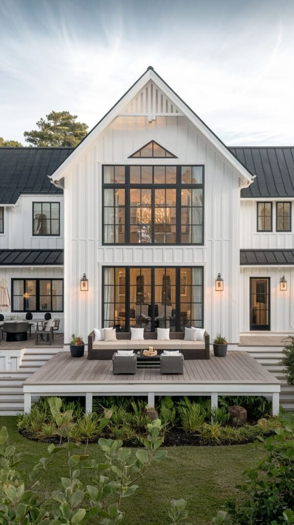 Rear view of a large white farmhouse with a black metal roof. The central two-story gable window wall overlooks a tiered, dark wood deck used for seating and entertaining.