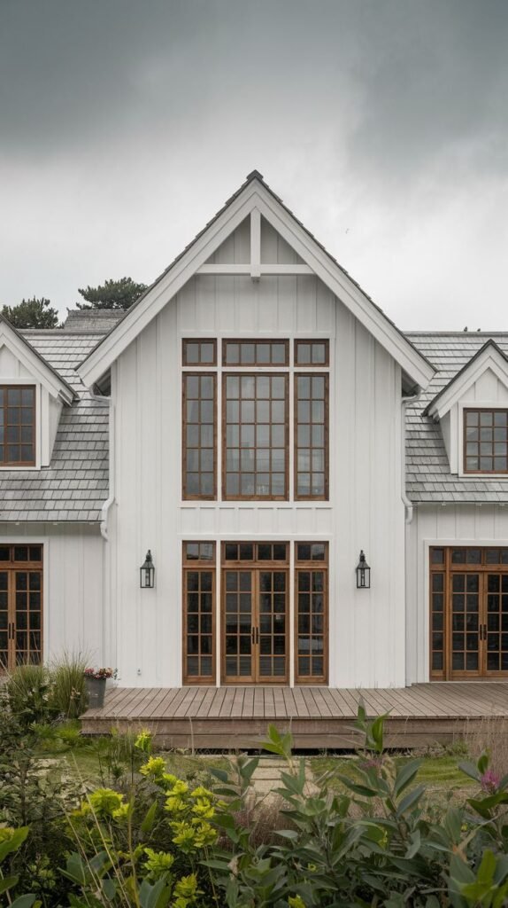 Symmetrical white farmhouse facade with a gray shingle roof and dormers. The central gable has a tall window grid with warm brown wood frames, matching the double entry doors and the deck flooring.