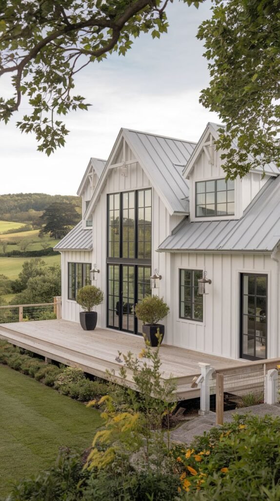 White modern farmhouse set on a hill, featuring a light metal roof and black-framed windows, including a large two-story central window wall. An expansive, elevated wood deck overlooks the green countryside.