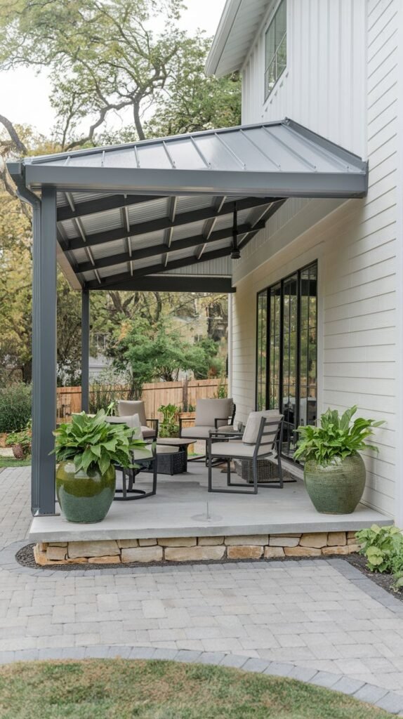 Small covered porch with a pitched metal roof, set on a low stacked stone base, attached to a white house, flanked by two large green ceramic planters.