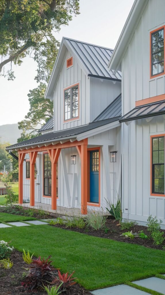 White board and batten farmhouse with a gray metal roof, featuring bold orange-red accent framing on the porch structure and window trim, plus a deep blue front door.