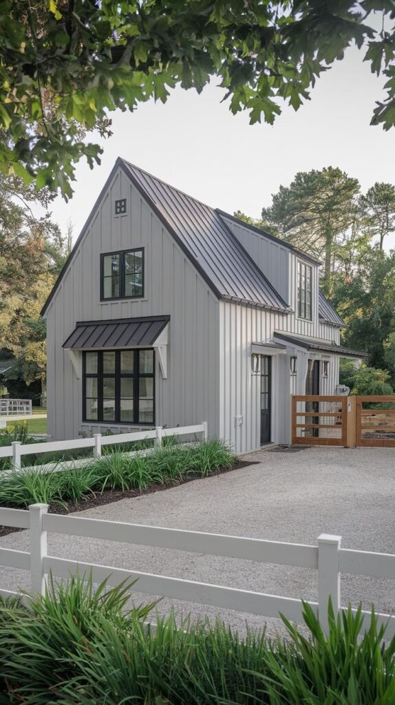 Light gray modern farmhouse structure with vertical siding, a dark metal roof, and high-contrast black window frames and door, surrounded by a white fence and gravel driveway.