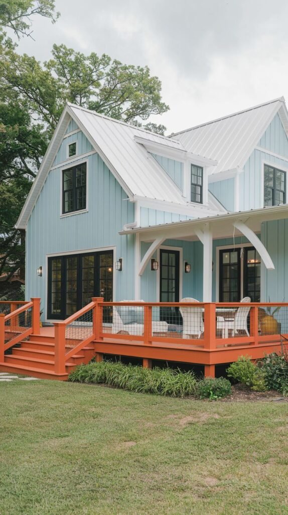 Light blue farmhouse with white metal roof and trim, featuring a large outdoor deck and stairs painted a vibrant orange/terra cotta color, complemented by black French doors.