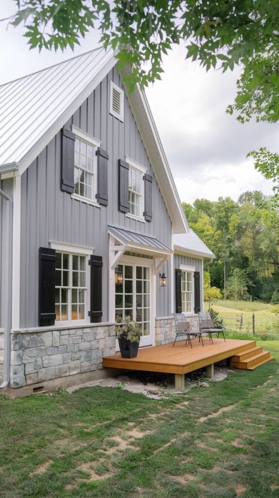 Gray vertical siding farmhouse above a rustic natural stone base, featuring black shutters, a white metal roof, and a wooden deck leading to French doors.