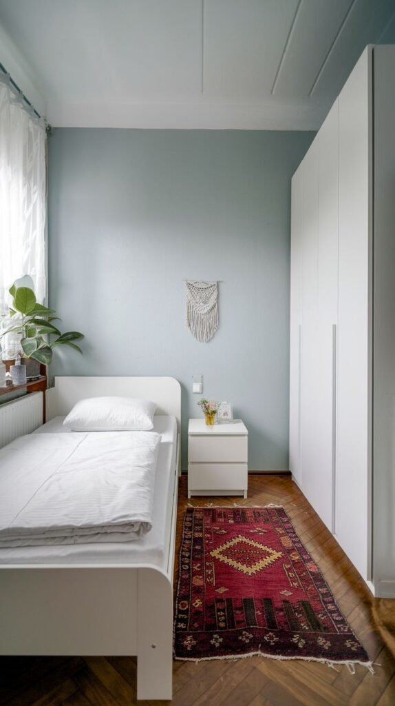 Modern Small Bedroom in a narrow space with pale blue walls, a large, seamless white wardrobe unit, and a small white bed centered on a contrasting dark red patterned rug.