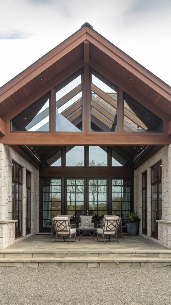 Formal covered entrance patio featuring a vaulted gable roof with a clerestory section filled with triangular glass windows, attached to a light stone house.