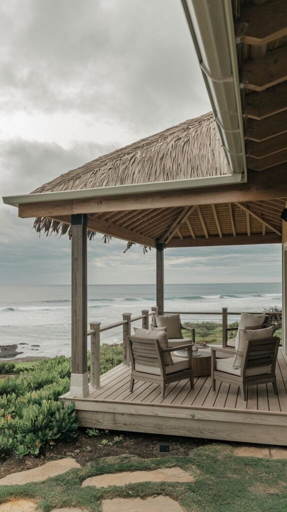 Elevated wooden deck with a tropical thatched roof supported by wood posts, offering a view of the ocean. The patio furniture is simple and set on the wooden flooring.