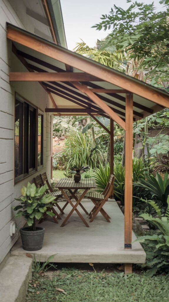 Concrete patio surrounded by dense tropical plants, covered by a simple, steeply sloping shed roof supported by wood. Features a folding wooden table and chairs.