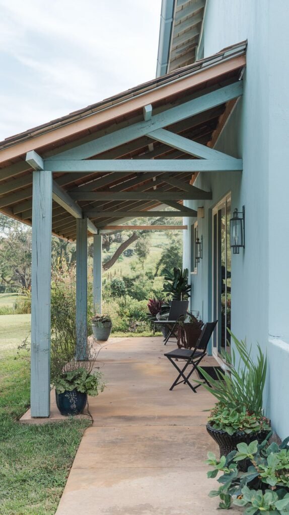Extended concrete patio running along a pale blue house wall. Covered by a long shed roof with exposed wooden beams and trusses, painted blue-gray.
