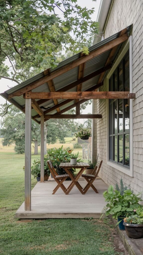 Elevated wood deck with a corrugated metal shed roof and rustic wood posts, overlooking a field. Furnished with a folding wooden table and chairs, situated against a shingle-sided house.