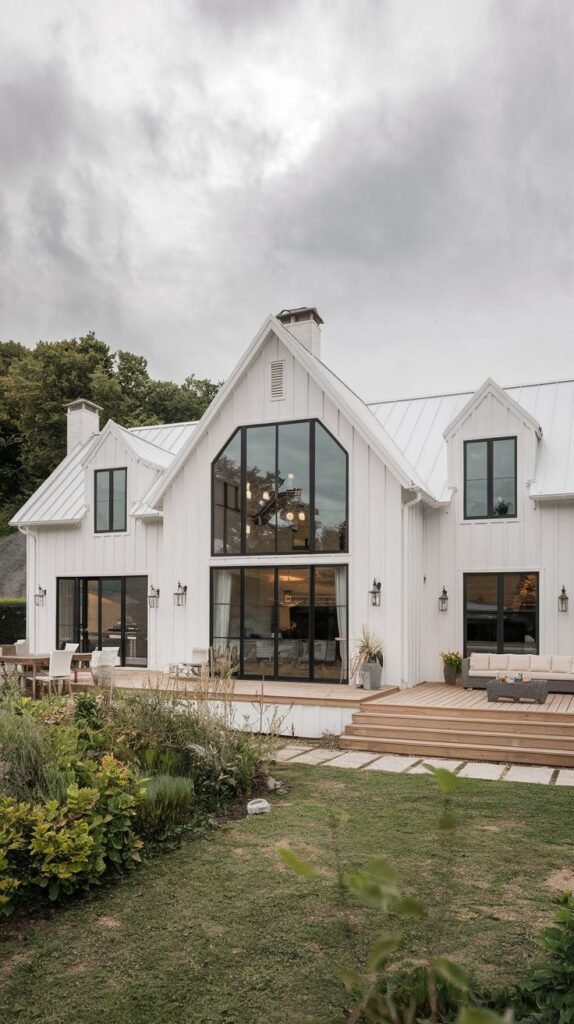 Rear view of a large, multi-gabled white farmhouse featuring an immense, custom triangular window wall in the central gable. A large light wood deck is integrated with stone pavers and patio furniture.