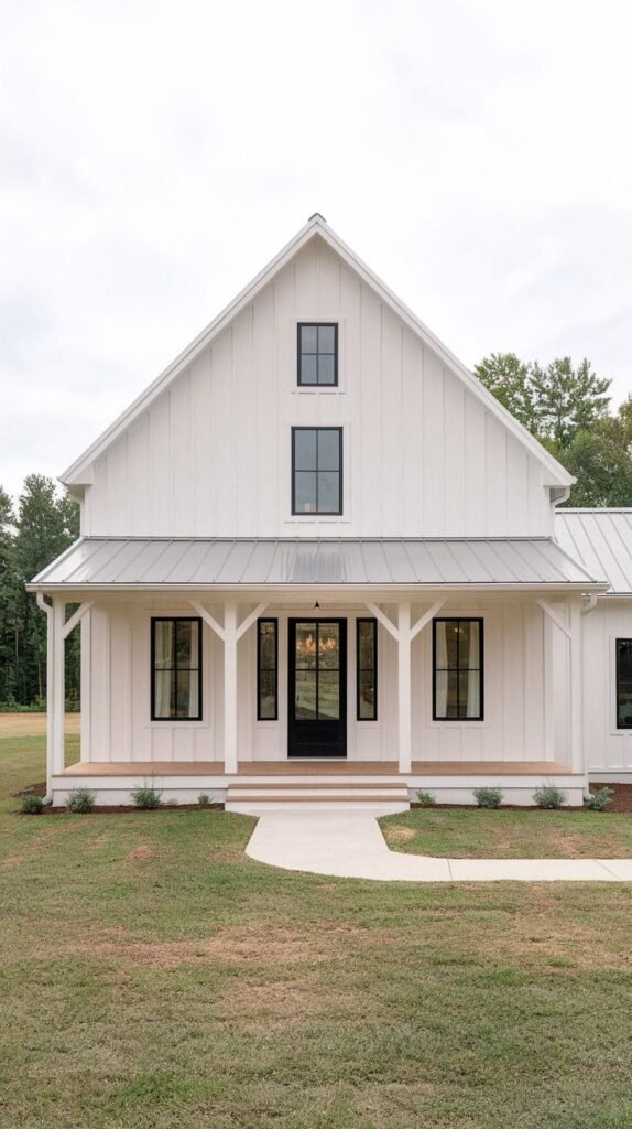 Front facade of a classic white farmhouse featuring a central covered entry porch with white tapered columns. The roof above the porch is light gray metal, and a curved concrete path leads to the steps.