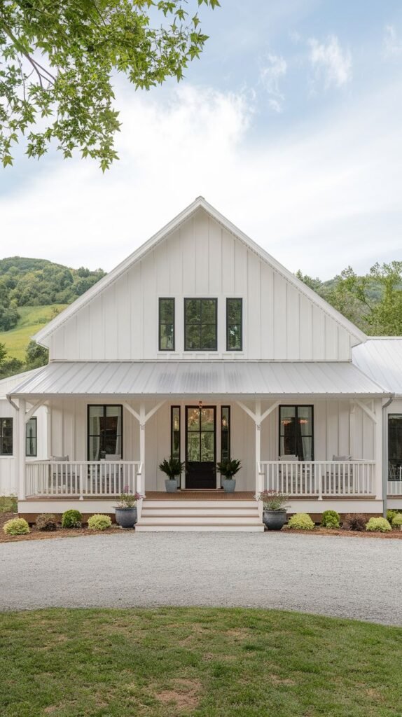 Traditional white farmhouse facade with a wide, covered front porch beneath a dark metal roof. The entrance is centered on the view, framed by white railings and dark porch columns, set against rolling green hills.