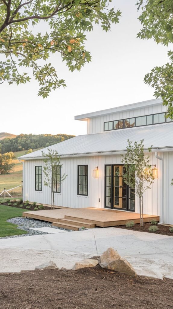 Side view of a white farmhouse addition with a low-slung white metal roof and black-framed windows. A light wood deck is attached, illuminated by modern black sconces, set against green rolling hills.
